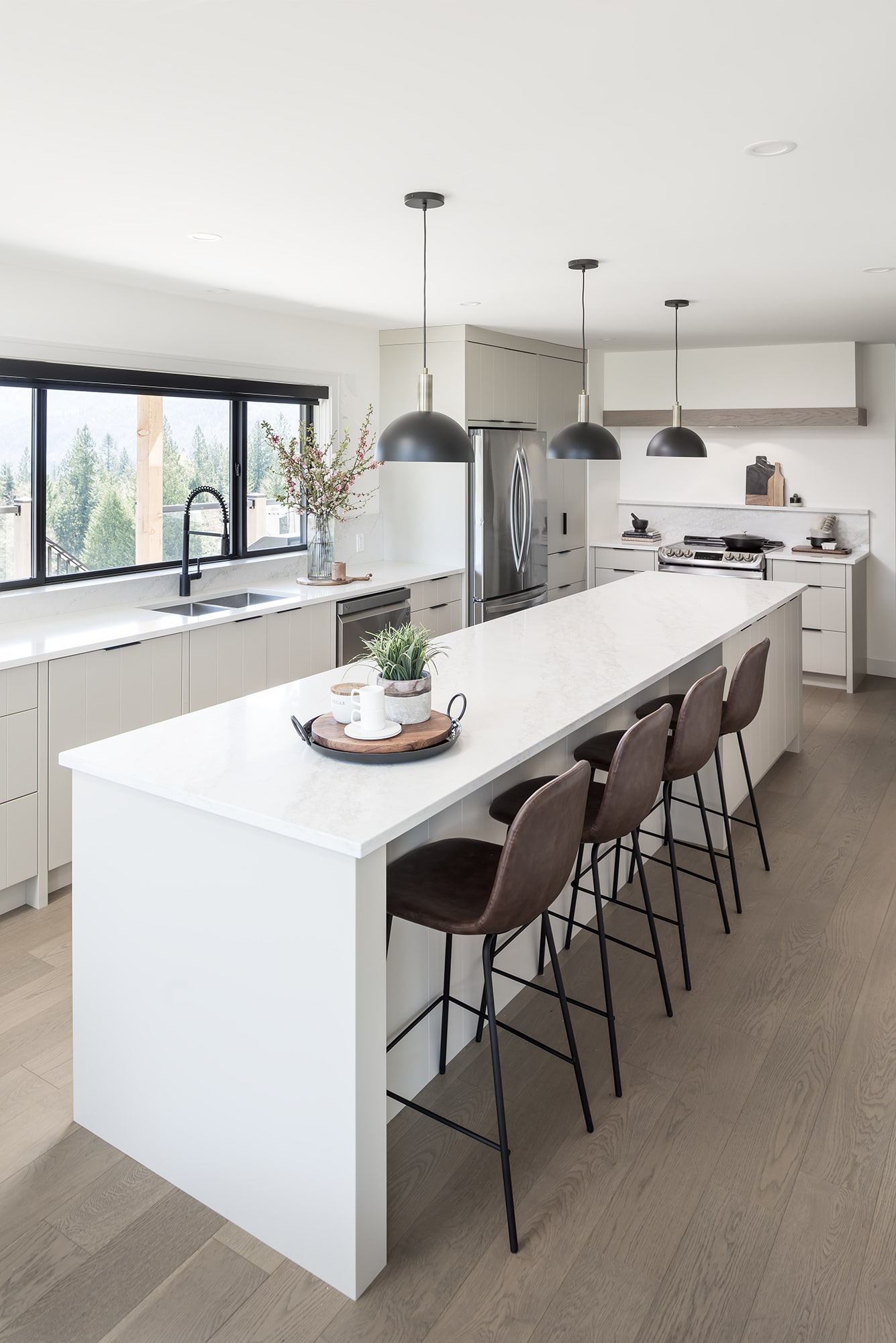 Modern all-white kitchen with custom waterfall island and cabinetry by Alderidge Construction, featuring black bar stools, pendant lighting, and large windows