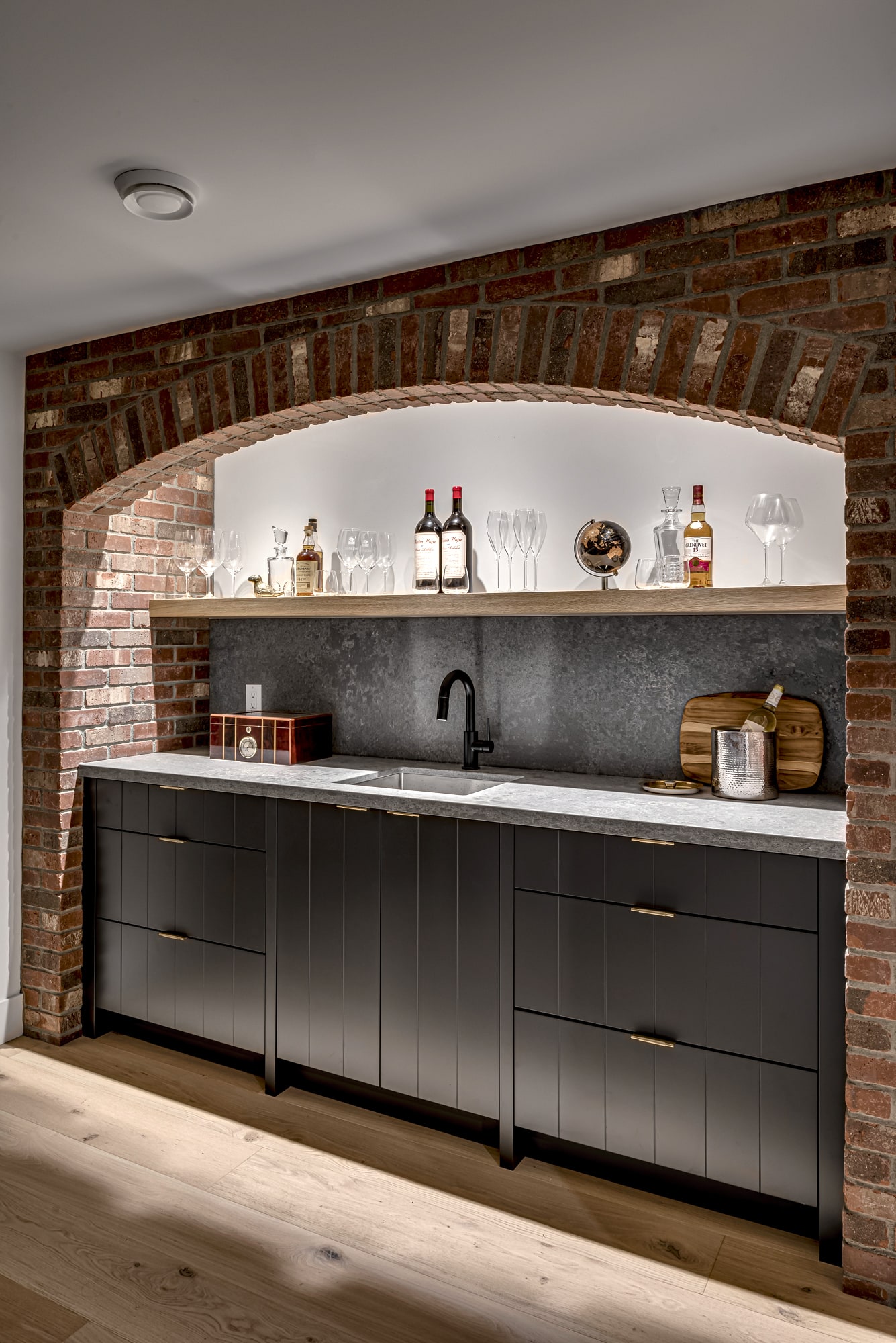 Custom dark cabinetry bar with brick arched alcove and open shelving by Alderidge Construction, showcasing dramatic custom millwork and built-in cabinetry design