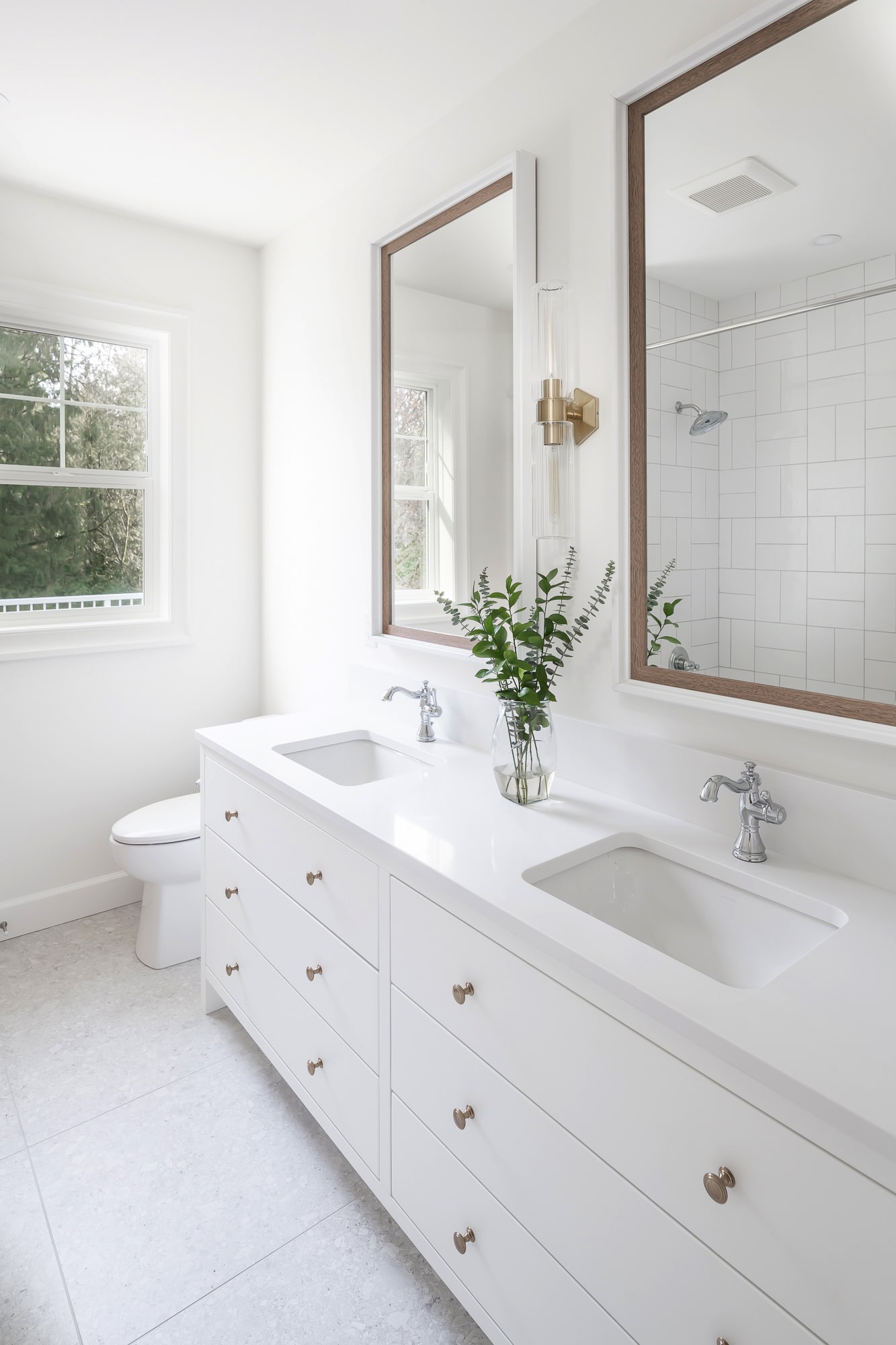 Custom bathroom with white vanity cabinetry by Alderidge Construction, featuring a wood-framed mirror, gold wall sconce, and bright white tile shower surround