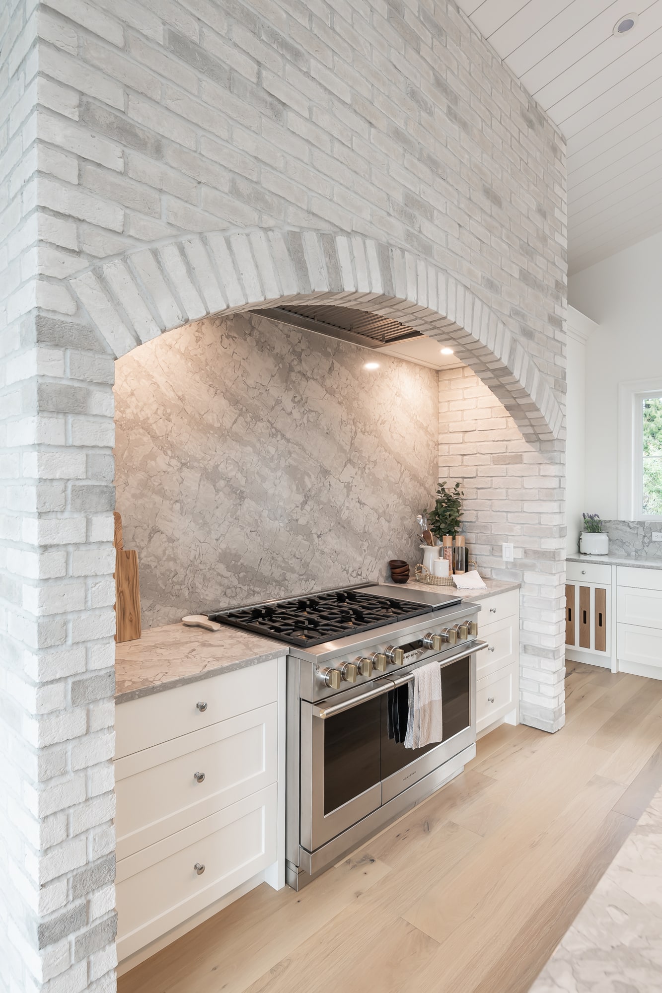 Custom kitchen with arched brick range hood surround and white cabinetry by Alderidge Construction, featuring marble slab backsplash and professional gas range