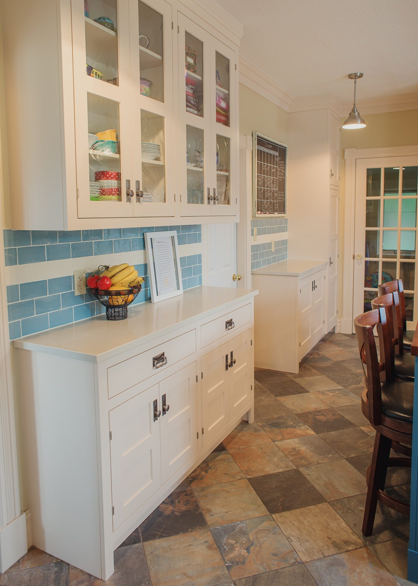 Custom white glass-front upper kitchen cabinetry by Alderidge Construction with blue subway tile backsplash, farmhouse sink, and vintage pendant lighting