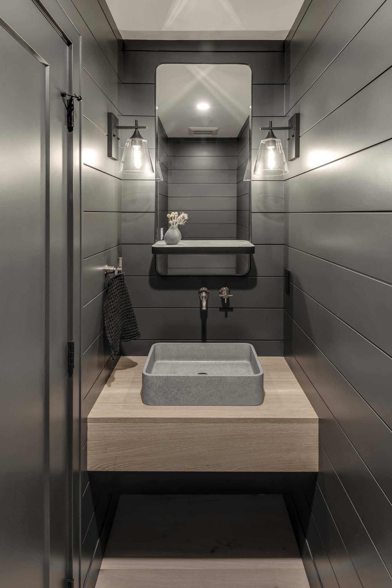 Custom floating powder room vanity with natural wood cabinetry by Alderidge Construction, featuring a concrete vessel sink, matte black faucet, and dark wall paneling